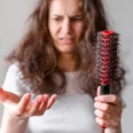 Worried woman holding a hairbrush in one hand and fallen hair in the other hand, concerned about hair fall and thin hair.